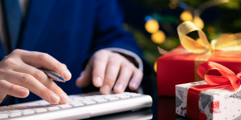 Mains d’un professionnel en costume écrivant sur clavier, avec des cadeaux de Noël en arrière-plan – image illustrant la préparation de fin d’année.